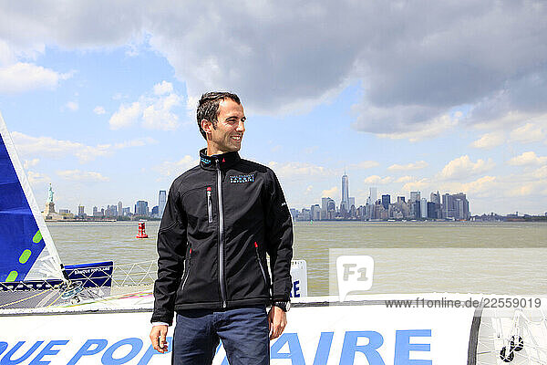 Man stands on the the Maxi Trimaran Solo Banque Populaire VII in front of the New York City skyline.