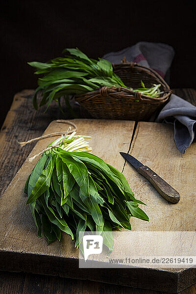 Bunch of wild garlic on a cutting board with a knife and willow basket on dark wood