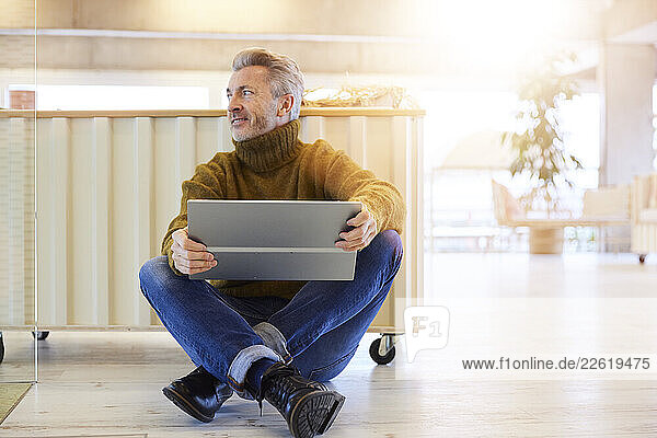 Mature man with digital tablet looking away while sitting on floor at home