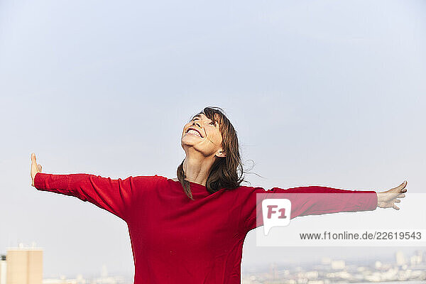 Mature woman looking up with arms outstretched on building terrace against clear sky