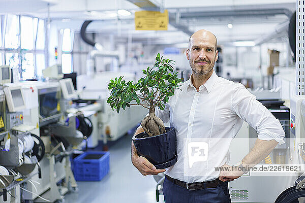 Confident male professional holding potted plant while standing with hand on hip at factory