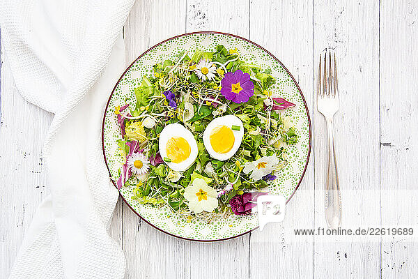 Spring salad with hard boiled eggs and edible flowers on a decorative plate with a fork on a white wooden table
