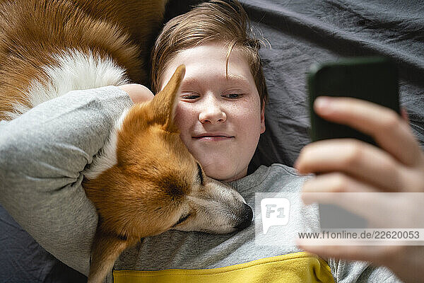 Portrait of boy lying on bed with his dog taking selfie with smartphone