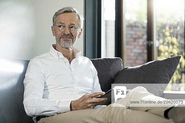 Senior man with grey hair in modern design living room sitting on couch holding tablet