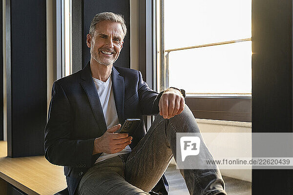 Smiling man in an office holding a mobile phone during sunset