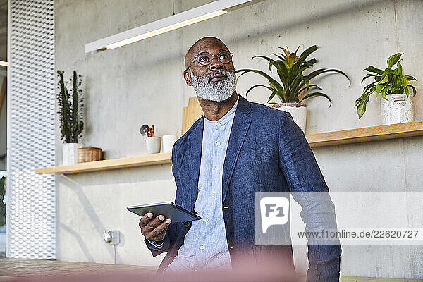 Mature man looking up while holding digital tablet standing at home