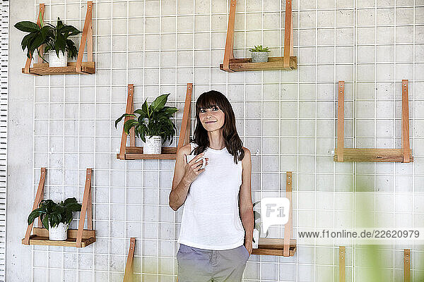 Mature woman with hands in pockets drinking coffee while standing at home