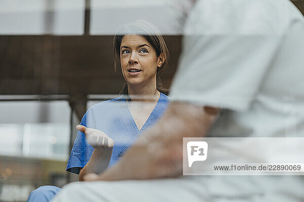 Female medical professional gesturing while discussing with coworker seen through glass in clinic