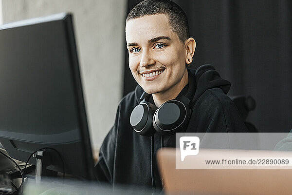 Portrait of smiling non-binary IT professional with wireless headphones around neck sitting in tech hub
