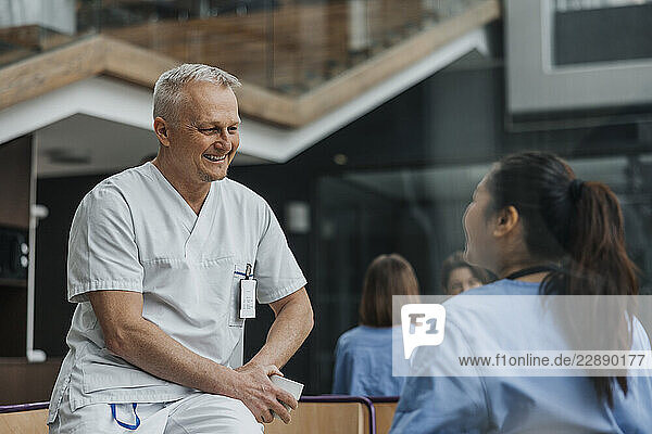 Happy mature male doctor talking with colleague while sitting on table in hospital cafeteria
