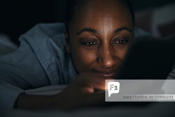 Smiling woman using smart phone while lying on bed in bedroom at night