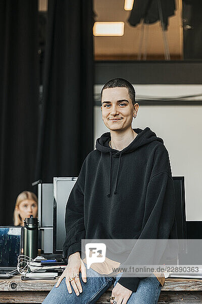 Portrait of smiling non-binary IT professional sitting on desk at work place