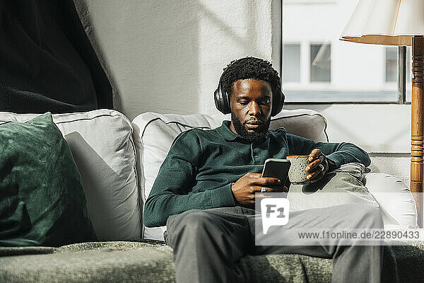 Young businessman listening to music through wireless headphones while using smart phone sitting on sofa at office