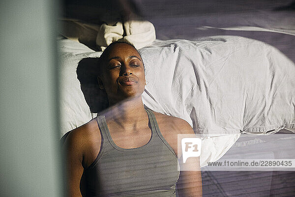Tired woman with eyes closed leaning on bed in bedroom seen through glass