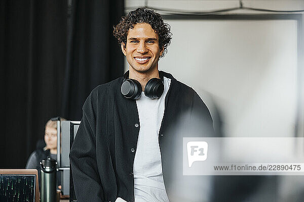 Portrait of smiling male computer programmer with wireless headphones around neck standing in tech hub