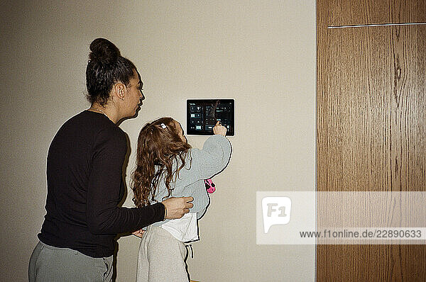 Mother assisting girl operating tablet PC mounted on wall at home