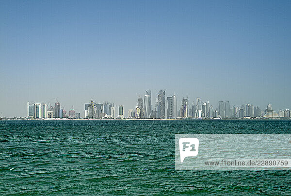 Skyscrapers of a modern city skyline reflected in the calm turquoise sea under a clear blue sky. Qatar