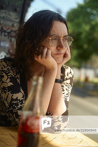 Pensive woman with glasses rests her chin on hands at an outdoor table with a soda bottle. Berlin  Germany