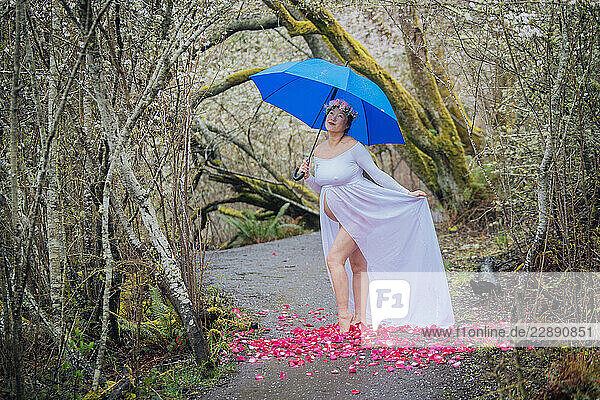 Pregnant woman in flowy white dress holds blue umbrella on forest path with pink flower petals. WA  USA