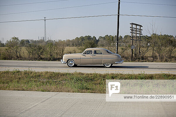 Vintage beige car driving on a rural road with a grassy field and trees in the background. Road Trip USA