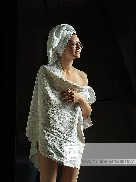Woman wrapped in a towel with a towel on her head against a dark background.