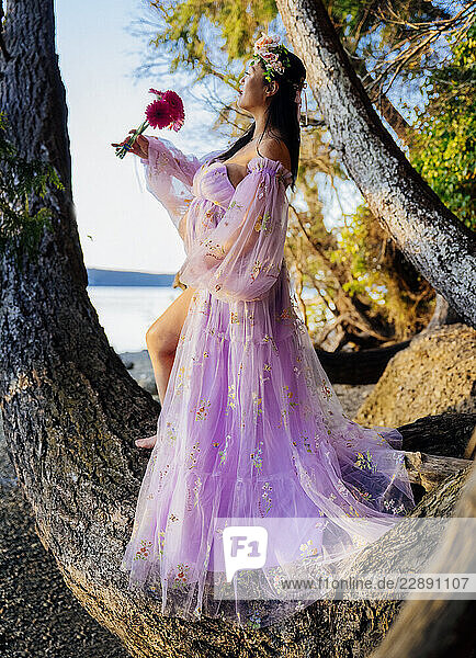 Pregant woman in a pink floral dress stands on tree branch by the sea with a flower. WA  USA