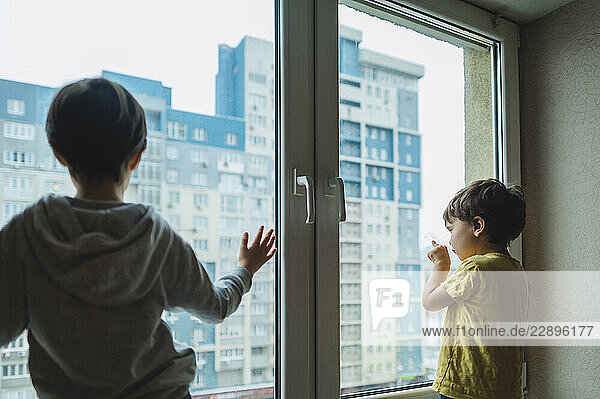 Two brothers looking out of a window at a cityscape from indoors during quarantine