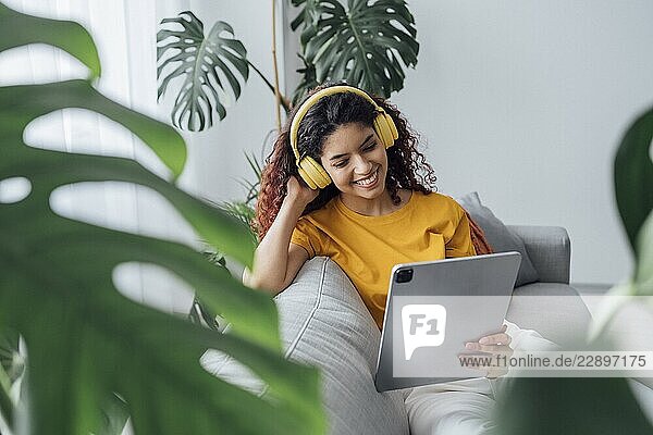 Woman relaxing on a sofa with headphones and a tablet surrounded by plants